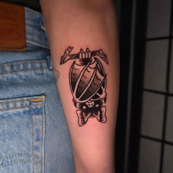 A bold black and grey traditional style tattoo of a bat hanging upside down on the forearm — by Miguel ANGELO, temecula, ca