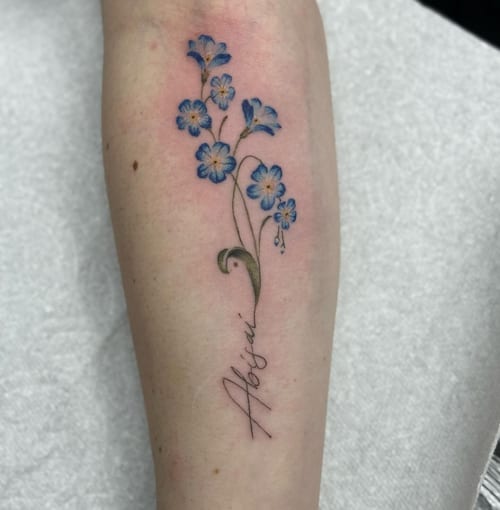 A delicate color tattoo of blue flowers with script lettering on the forearm, done in a fine line style — by Sandra Price, los angeles