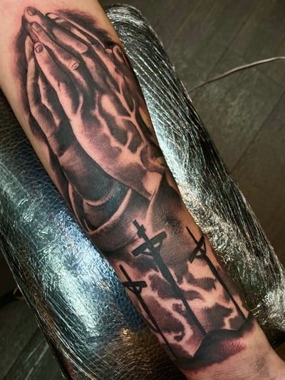 A black and grey tattoo on the forearm depicting realistic praying hands and crosses, symbolizing religious themes — by Josue Bravo, los angeles