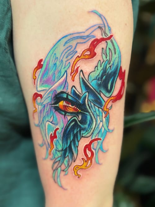 A vibrant neo-traditional tattoo of a stylized bird with flames on the upper arm, featuring bold colors and dynamic lines — by Tweetwee, los angeles