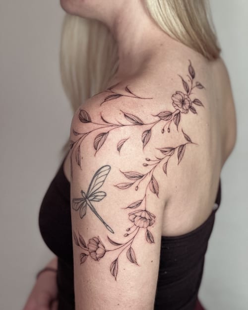 A delicate black and grey tattoo of a dragonfly and floral vine on the shoulder, featuring fine line work and minimalistic design — by Melanie Benoit, los angeles