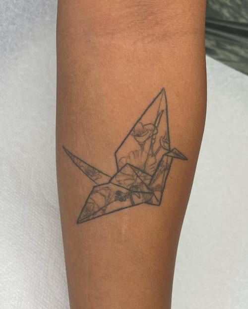 A delicate black and grey origami crane tattoo on the forearm with fine line floral details — by Sandra Price, los angeles