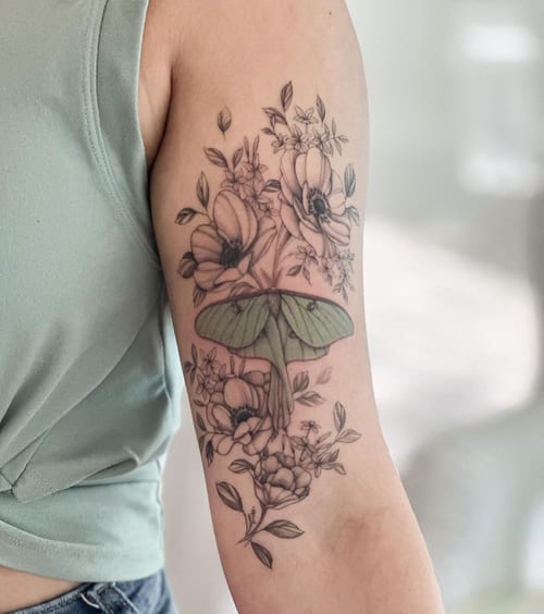 A delicate tattoo of a moth surrounded by flowers on the upper arm, featuring fine line work and a mix of black and grey with color accents — by Melanie Benoit, los angeles