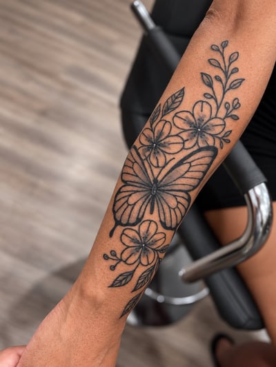 A delicate black and grey butterfly and floral tattoo on the forearm, featuring fine line work and clean details — by HEAVY INK, los angeles
