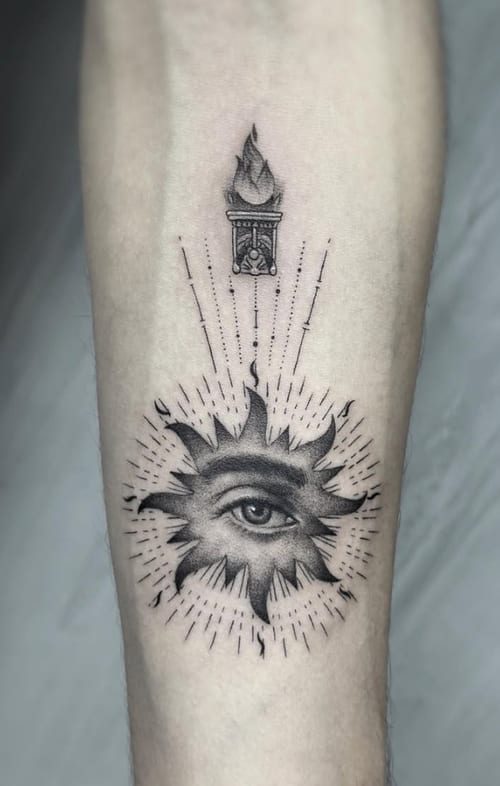 A black and grey surrealistic tattoo on the forearm featuring an eye within a sunburst design, topped with a stylized flame — by Jian Liang, los angeles