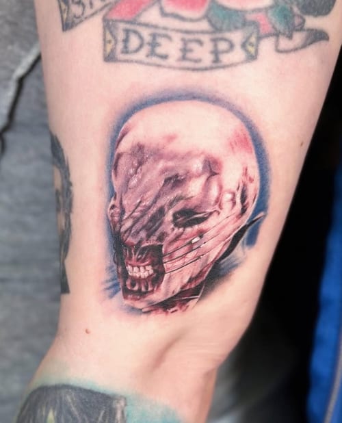 A horror-themed tattoo of a distorted skull with realistic details and a mix of color and black ink on the forearm — by Jian Liang, los angeles