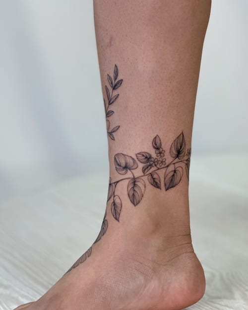 A delicate black and grey floral vine tattoo wrapping around the ankle, featuring fine line details — by Melanie Benoit, los angeles