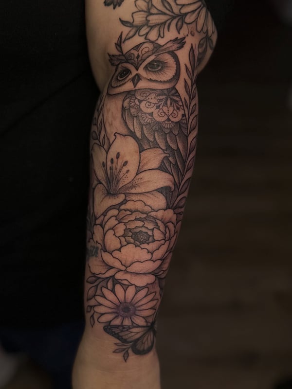 A delicate black and grey owl and floral tattoo on the forearm — by Maddie Ramirez, palm desert, ca