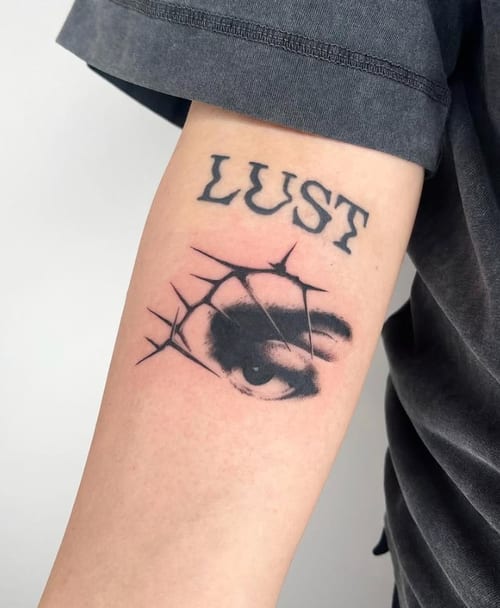 A black and grey tattoo of an eye with thorn-like lines and the word 'LUST' above it on the upper arm — by Level tattoo, los angeles