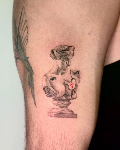 A black and grey tattoo of a bust sculpture with script text on the thigh, featuring a small red heart for contrast — by zealous, los angeles