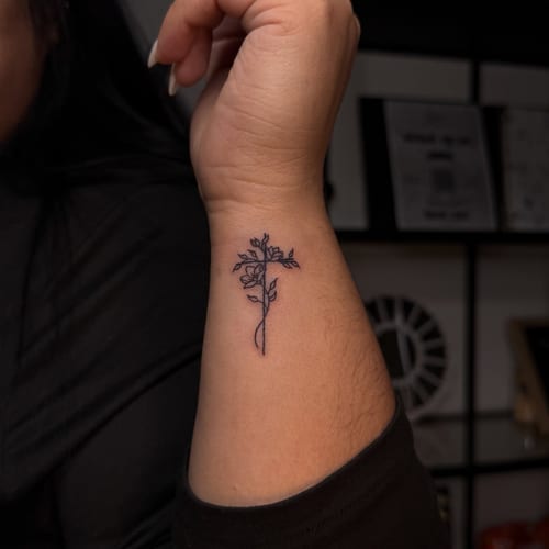 A delicate fine line flower tattoo on the wrist, featuring a minimalist black and grey design — by NJ TATTOOZ, los angeles