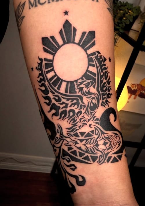 A black and grey tribal-style tattoo on the forearm featuring a stylized sun and a lion with bold lines and geometric patterns — by NJ TATTOOZ, los angeles