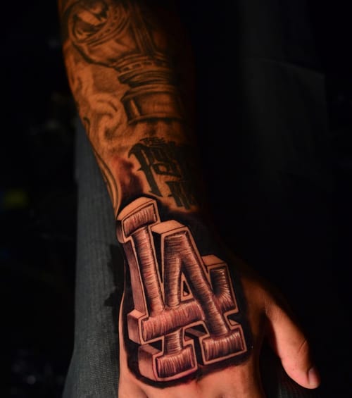 A bold black and grey LA lettering tattoo on the hand with strong shading and script style — by Johnny Ayala, los angeles