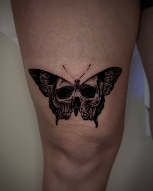 A black and grey surrealistic tattoo of a butterfly with skulls integrated into its wings on the thigh — by emily, rahway
