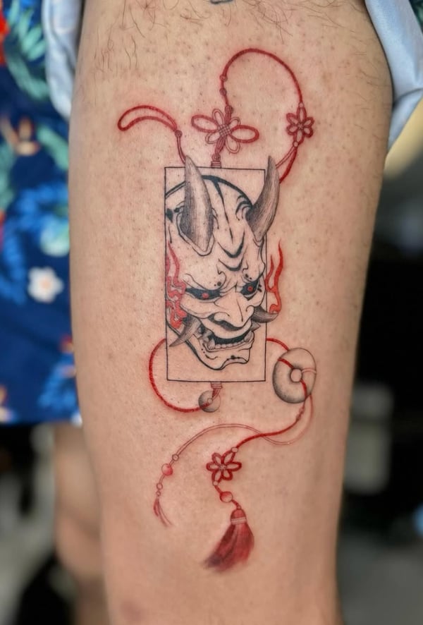 A mixed color and black Japanese-style tattoo of a demon mask on the thigh, featuring red decorative elements and fine line details — by otaybeatdown, orange, ca