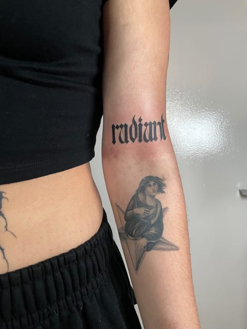 A bold black and grey forearm tattoo featuring the word 'radiant' in script lettering and a realistic figure sitting on a star — by Brian Kies, los angeles