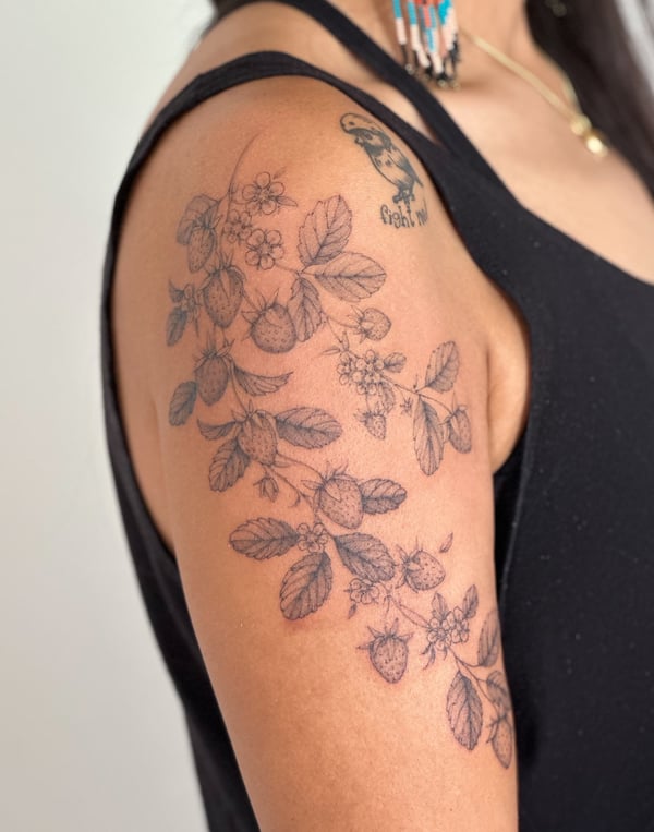 A delicate black and grey fine line tattoo of a strawberry plant with flowers on the upper arm — by Melanie Benoit, los angeles