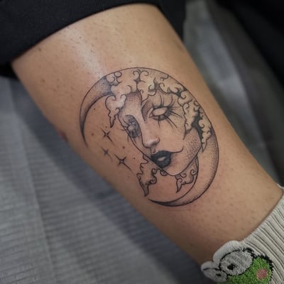 A delicate blackwork tattoo of a moon with a woman's face on the thigh, featuring dotwork shading and surreal elements — by Maddie Ramirez, los angeles