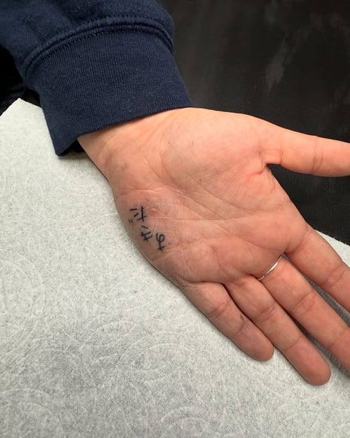 A minimalist black script lettering tattoo on the hand, featuring simple and clean lines — by zealous, los angeles