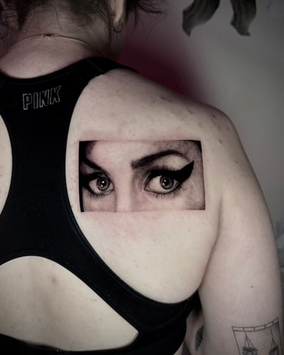 A black and grey realism tattoo of a woman's eyes and eyebrows on the upper back, featuring detailed shading and fine lines — by emily, los angeles