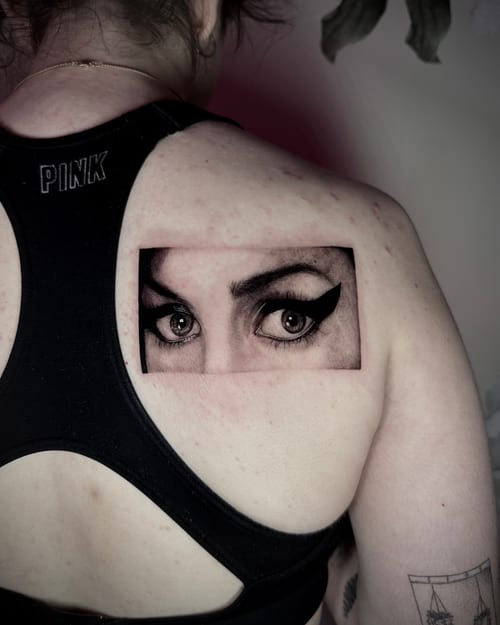 A black and grey realism tattoo of a woman's eyes and eyebrows on the upper back, featuring detailed shading and fine lines — by emily, los angeles