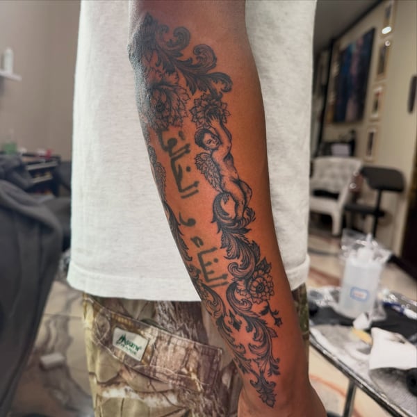 A detailed black and grey tattoo on the forearm featuring an ornamental design with a cherub and script lettering — by Kiera Garvin, los angeles