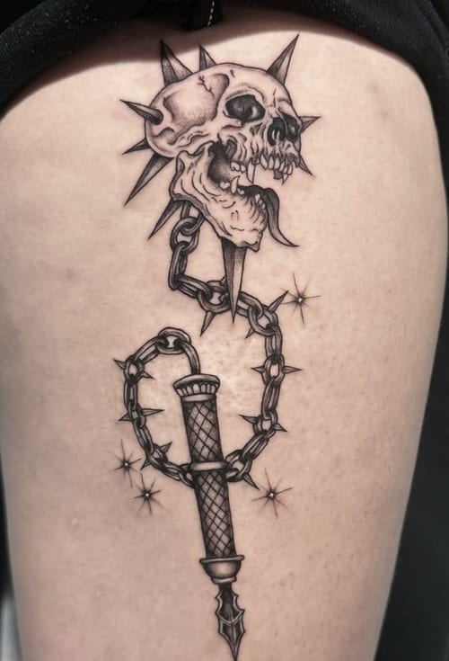 A black and grey illustrative tattoo on the thigh featuring a skull with a spiked chain and dagger, rendered in a bold blackwork style — by Shelby Bianco, los angeles