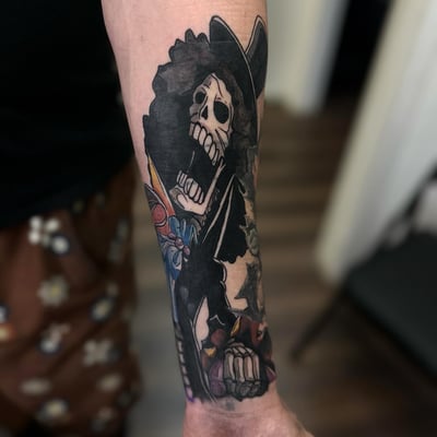 A colorful skeleton with an afro and hat tattoo on the forearm in a blackwork and illustrative style — by Maddie Ramirez, los angeles