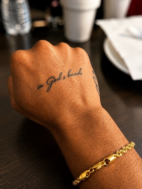 A minimalist black script lettering tattoo on the hand reading 'in God's hands' — by HEAVY INK, chino, ca