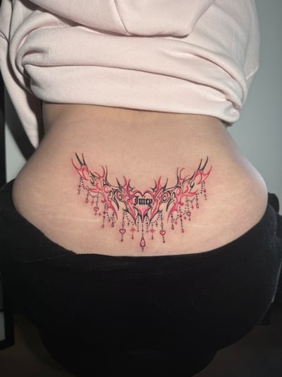An ornamental tattoo on the lower back featuring the word 'Juicy' in script with decorative elements and pink accents — by Shelby Bianco, los angeles