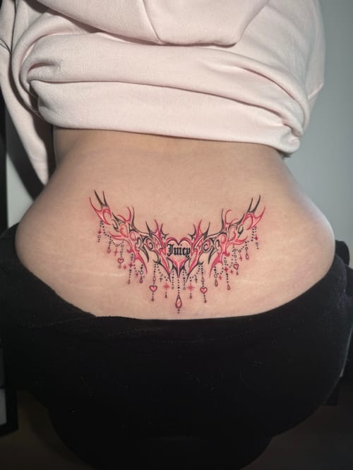 An ornamental tattoo on the lower back featuring the word 'Juicy' in script with decorative elements and pink accents — by Shelby Bianco, los angeles