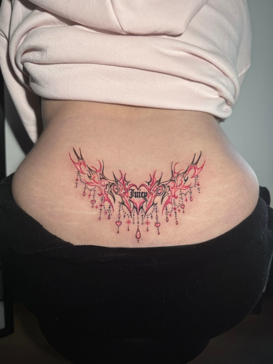 An ornamental tattoo on the lower back featuring the word 'Juicy' in script with decorative elements and pink accents — by Shelby Bianco, los angeles