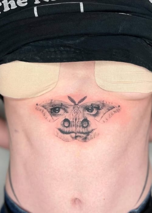 A surreal black and grey tattoo of a butterfly with human eyes on its wings, located on the stomach — by Level tattoo, los angeles