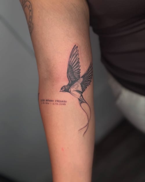 A delicate black and grey tattoo of a flying bird on the forearm, accompanied by memorial text — by zealous, los angeles