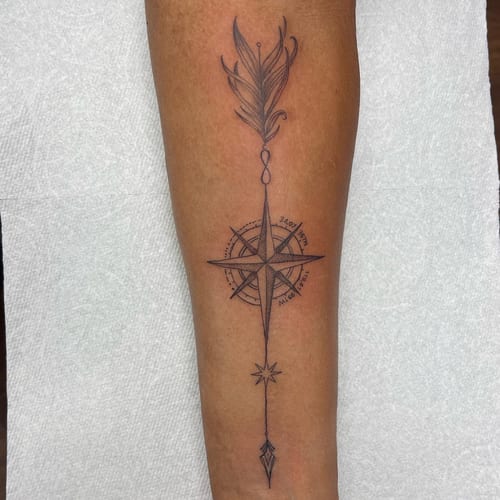 A delicate black and grey compass with a feather design on the forearm, featuring fine line and geometric elements — by Sandra Price, los angeles