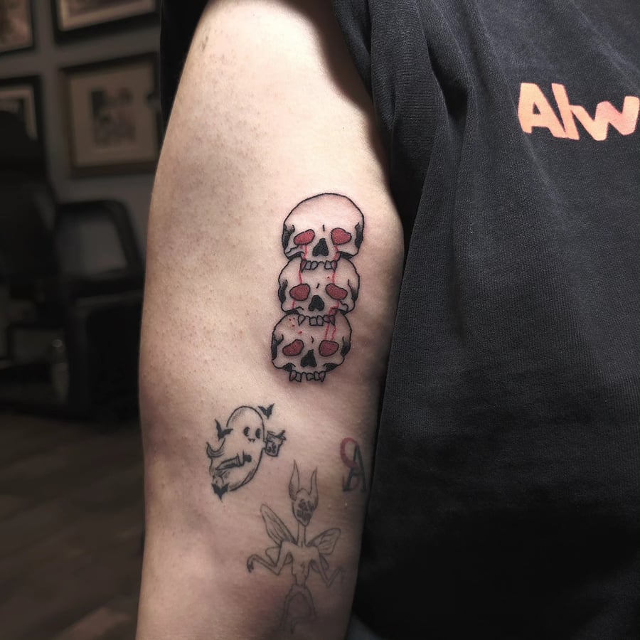 A bold and graphic stack of skulls tattoo on the upper arm, featuring traditional and illustrative styles with red accents — by Miguel ANGELO, los angeles