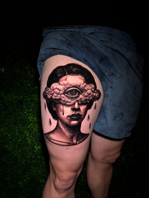 A surreal black and grey tattoo on the thigh depicting a woman's face with an eye in the forehead, surrounded by clouds and tears falling down — by Travis Ross, los angeles