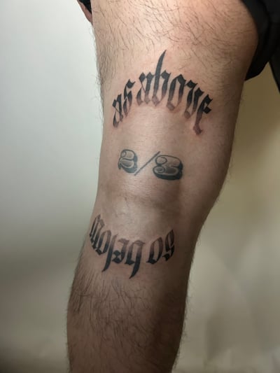 A black and grey lettering tattoo on the leg featuring script text with a bold, gothic style — by Brian Kies, los angeles