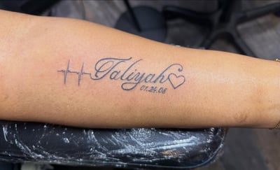 A minimalist black and grey tattoo featuring a name, heart, and date on the forearm in script lettering — by Kiera Garvin, los angeles