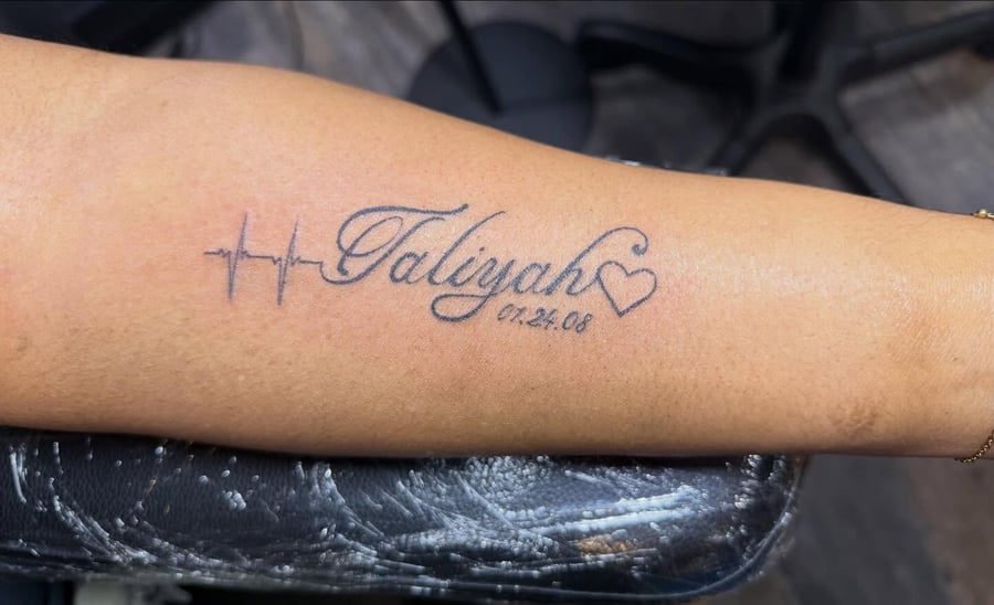 A minimalist black and grey tattoo featuring a name, heart, and date on the forearm in script lettering — by Kiera Garvin, los angeles