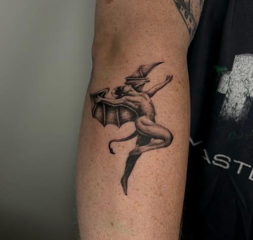A black and grey tattoo of a demon-like figure with wings and a hat on the forearm, rendered in a realistic style with detailed shading — by velvet devil, los angeles