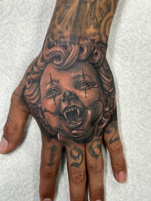 A black and grey realism tattoo of a child's face with clown makeup on the hand, featuring detailed shading and expression — by Humilde tattoos, los angeles