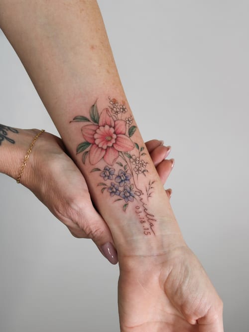 A delicate floral bouquet tattoo on the forearm with fine line details, featuring colorful flowers and a name with a date — by Melanie Benoit, los angeles