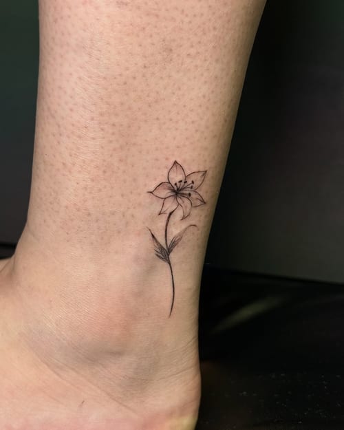 A delicate fine line flower tattoo on the ankle, featuring a minimalist black and grey design — by zealous, los angeles