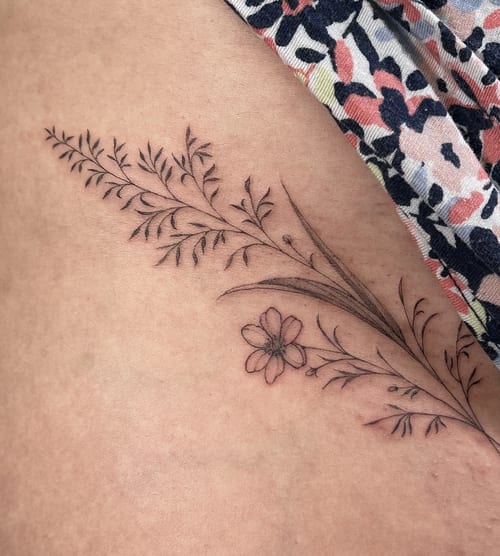A delicate black and grey fine line floral design tattooed on the hip, featuring minimalistic flower and leaf elements — by Melanie Benoit, los angeles