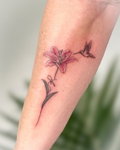 A delicate tattoo of a hummingbird hovering near a pink lily on the forearm, done in fine line style with a soft color palette — by Melanie Benoit, los angeles