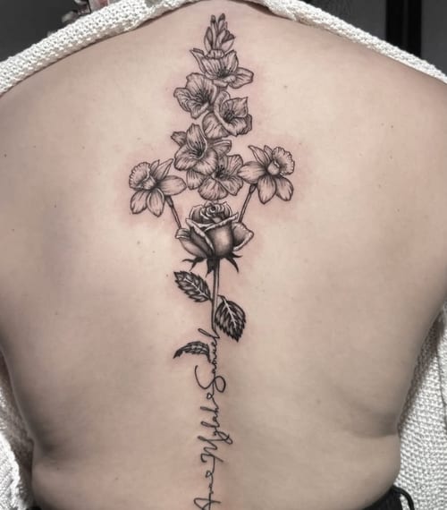A black and grey fine line tattoo of a floral arrangement with a rose and other flowers on the back, accompanied by script text — by Shelby Bianco, los angeles