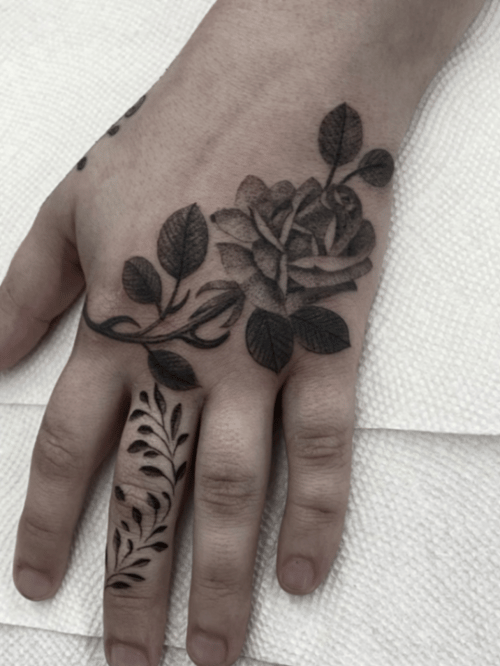 A black and grey floral tattoo on the hand featuring a rose and leaves with intricate detailing — by tyler_whitlock, los angeles