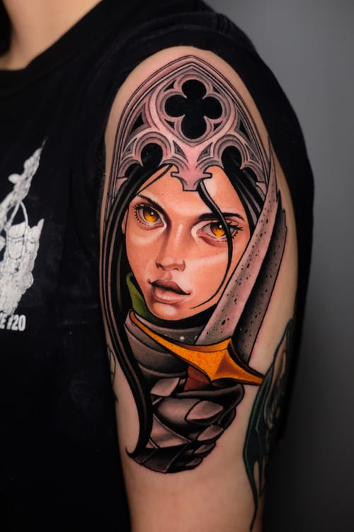 A vibrant neo-traditional tattoo on the upper arm depicting a woman's face with striking eyes, framed by a decorative helmet and holding a sword, featuring bold lines and rich colors — by Gabe Mott…