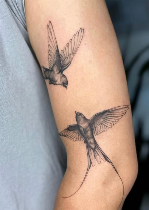 A black and grey fine line tattoo of two birds in flight on the upper arm, showcasing delicate linework and shading — by otaybeatdown, los angeles
